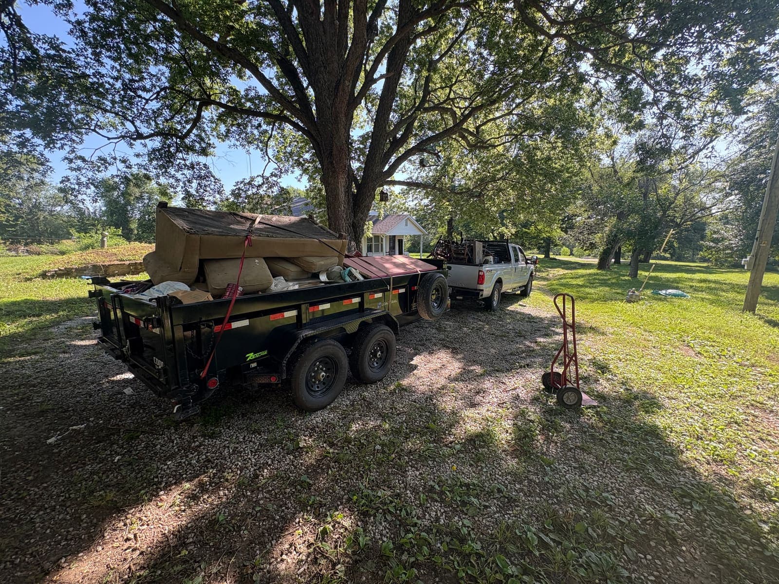 Dump trailer loaded with debris under a large oak at a farmhouse